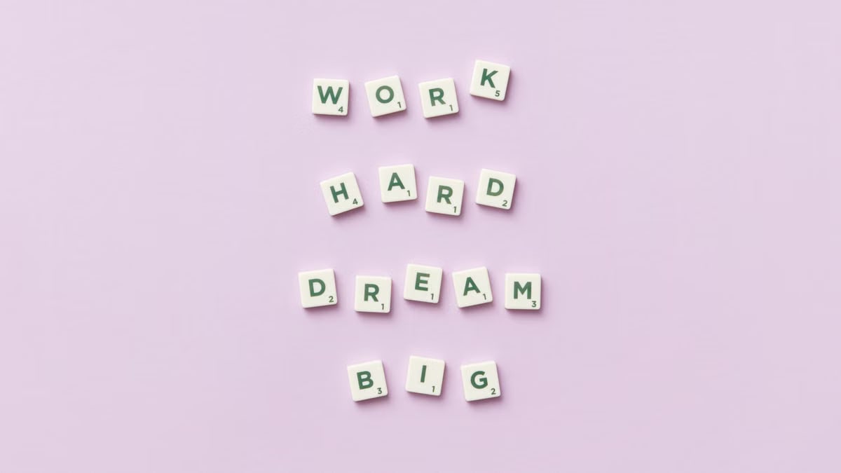 Scrabble tiles spell 'Work Hard Dream Big' on a pastel purple surface, top view.